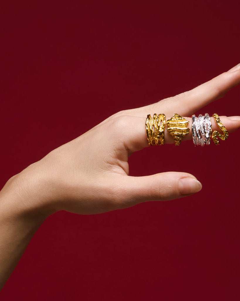 A woman in a white tank top, adorned with multiple 18K gold plated rings on her fingers, smiling softly at the camera.