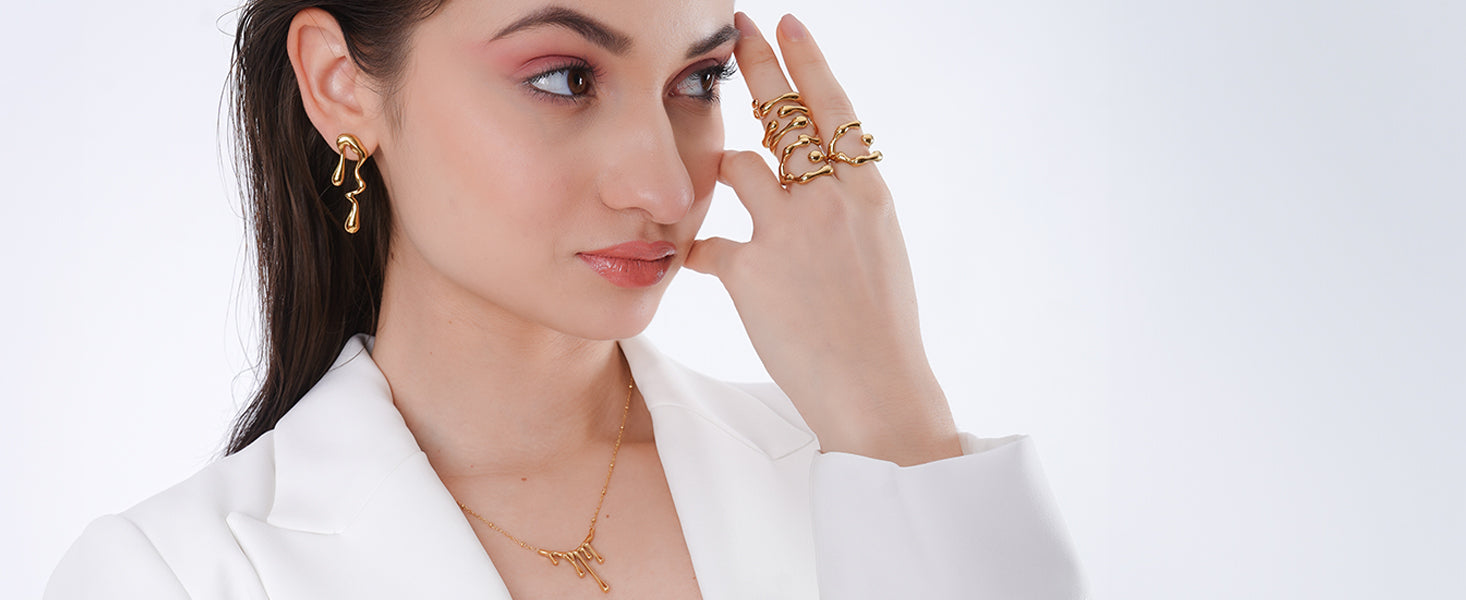 A woman in a white blazer showcases contemporary 18K gold-plated jewelry, including sculptural drop earrings and an intricate multi-finger ring featuring fluid, organic shapes with a blue accent. Her sleek, wet-look hairstyle and soft pink makeup complete the modern, refined aesthetic.
