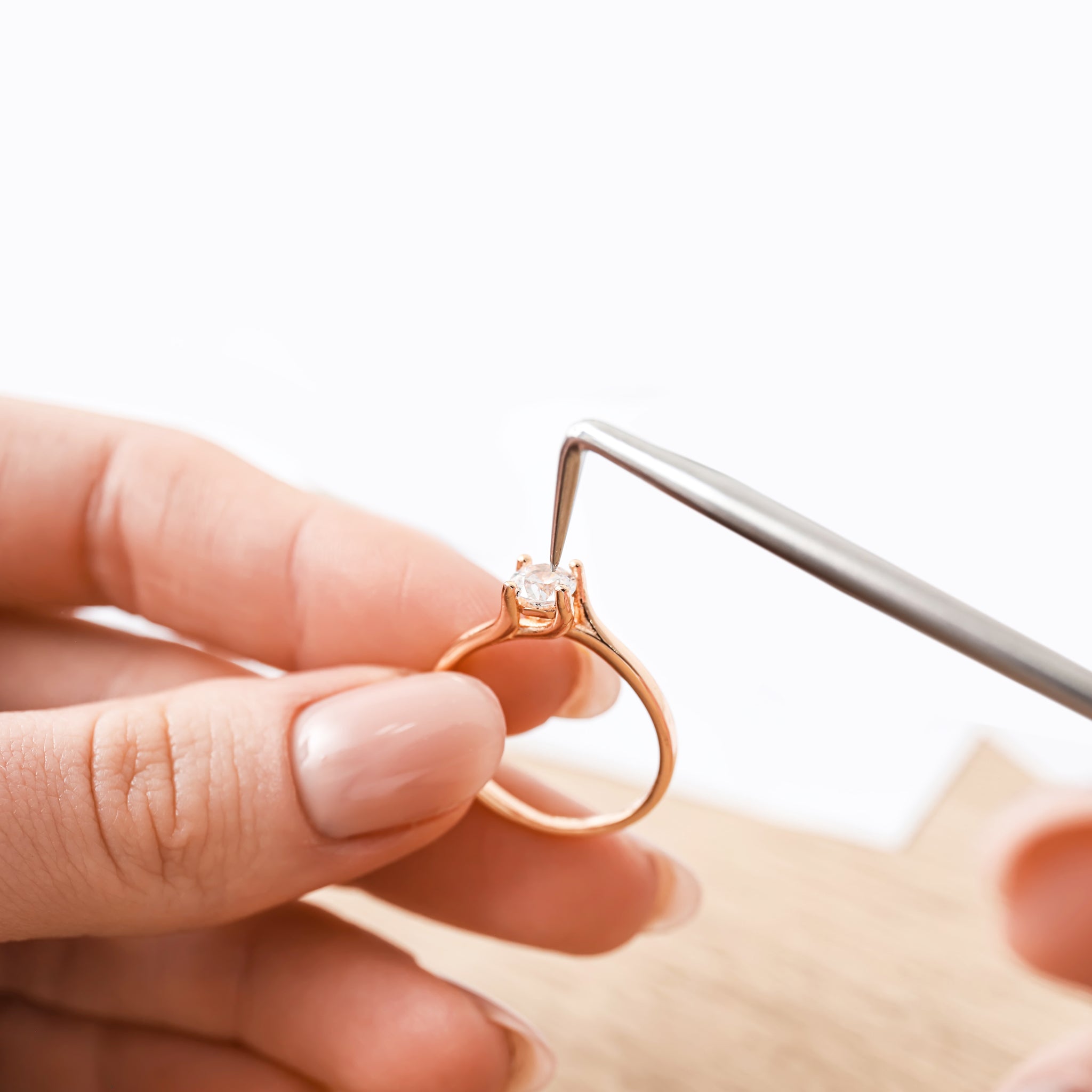 A close-up of a jeweler's hands working on a gold engagement ring with a round-cut diamond. One hand holds the ring, while the other uses fine-tipped tweezers to adjust the gemstone setting. The background is clean and minimalistic, emphasizing the craftsmanship and elegance of the jewelry.