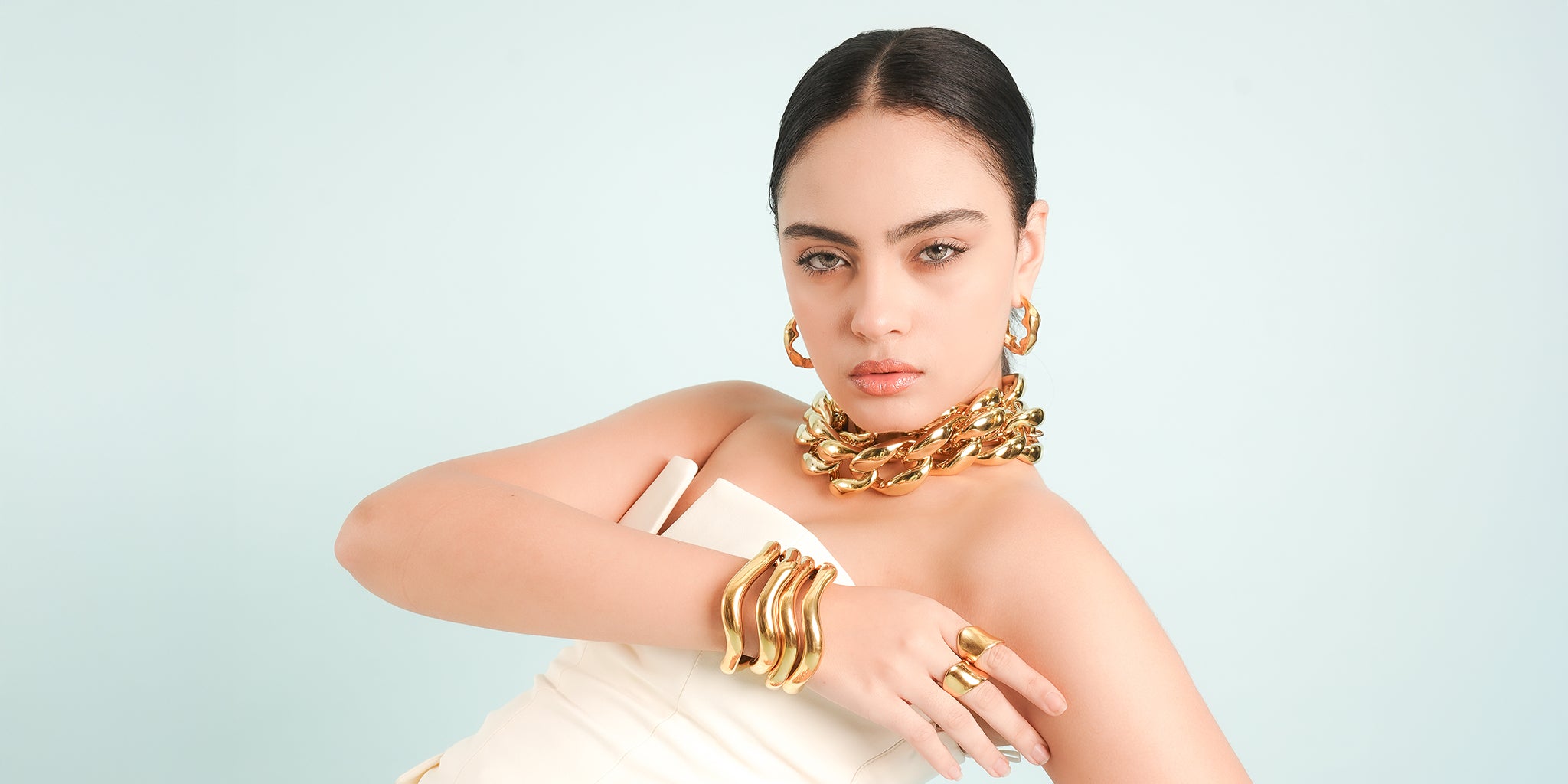 A woman adorned in bold 18K gold-plated jewelry, featuring a chunky chain necklace, sculptural hoop earrings, wavy cuff bracelets, and statement rings. Styled in an elegant white ensemble against a soft pastel backdrop, she exudes confidence and modern luxury.