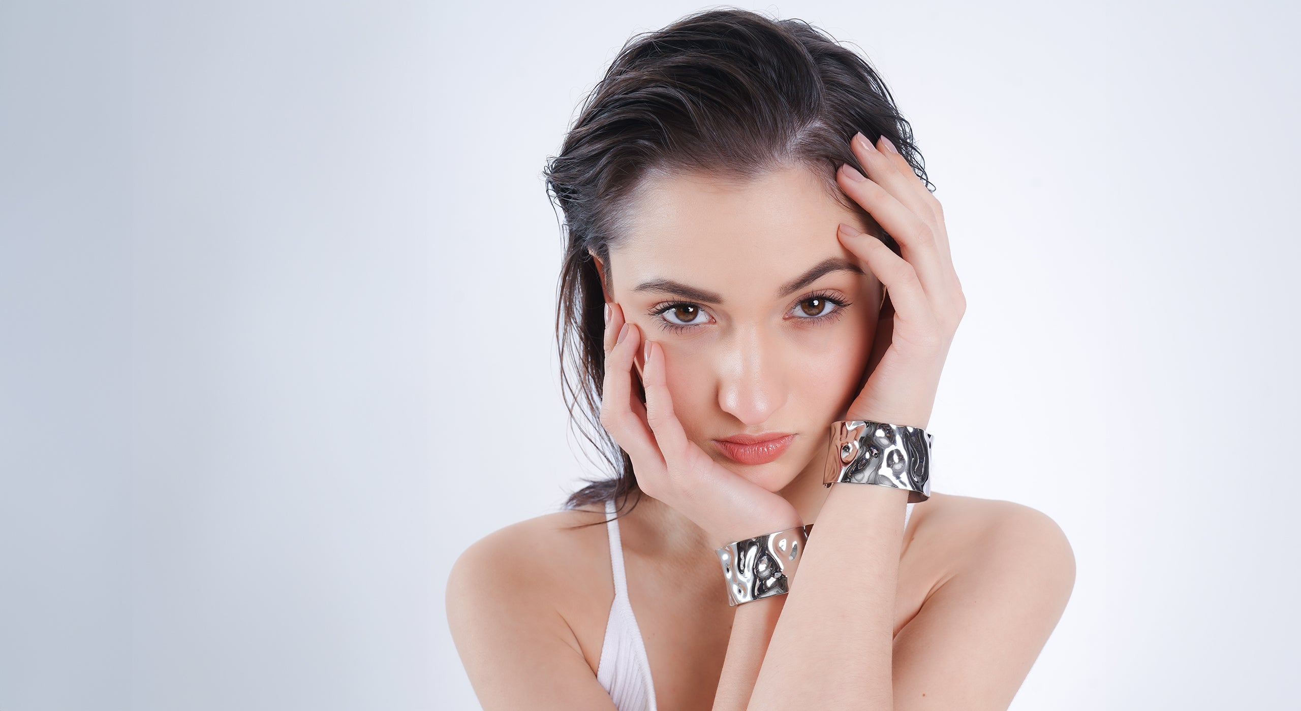 A woman with wet, slicked-back hair poses with her hands framing her face, showcasing bold, reflective silver cuff bracelets featuring a textured, wavy design. She wears a minimalist white outfit, exuding modern elegance and sophistication.