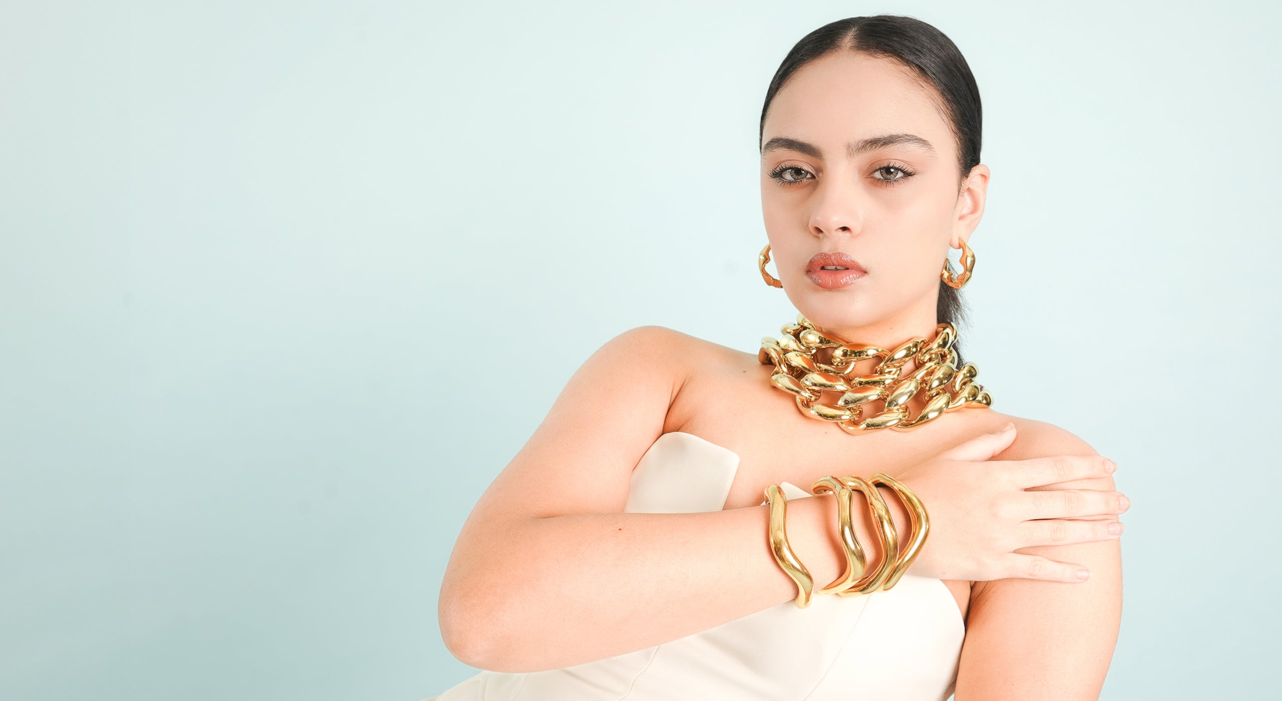 A woman in a strapless, elegant white outfit showcases bold gold accessories, including chunky chain necklaces, sculptural gold bracelets, and textured hoop earrings. The minimalist pastel background enhances the luxurious and statement jewelry.