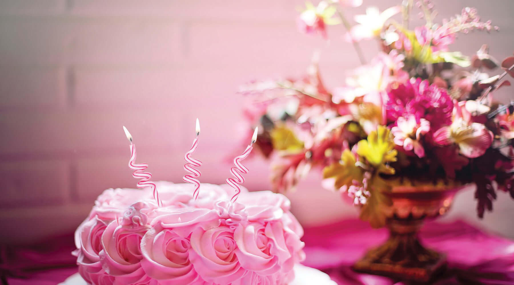 A beautifully decorated pink rosette cake with three spiral candles, placed on a white plate against a vibrant pink tablecloth, accompanied by a floral arrangement in the background.