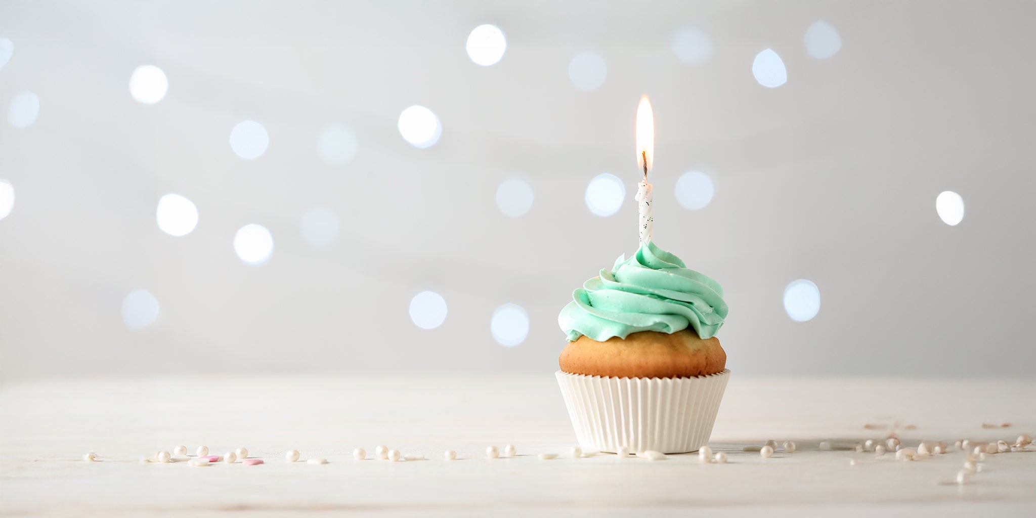 Cupcake with light blue frosting and a lit birthday candle, set against a soft, festive background with blurred lights.