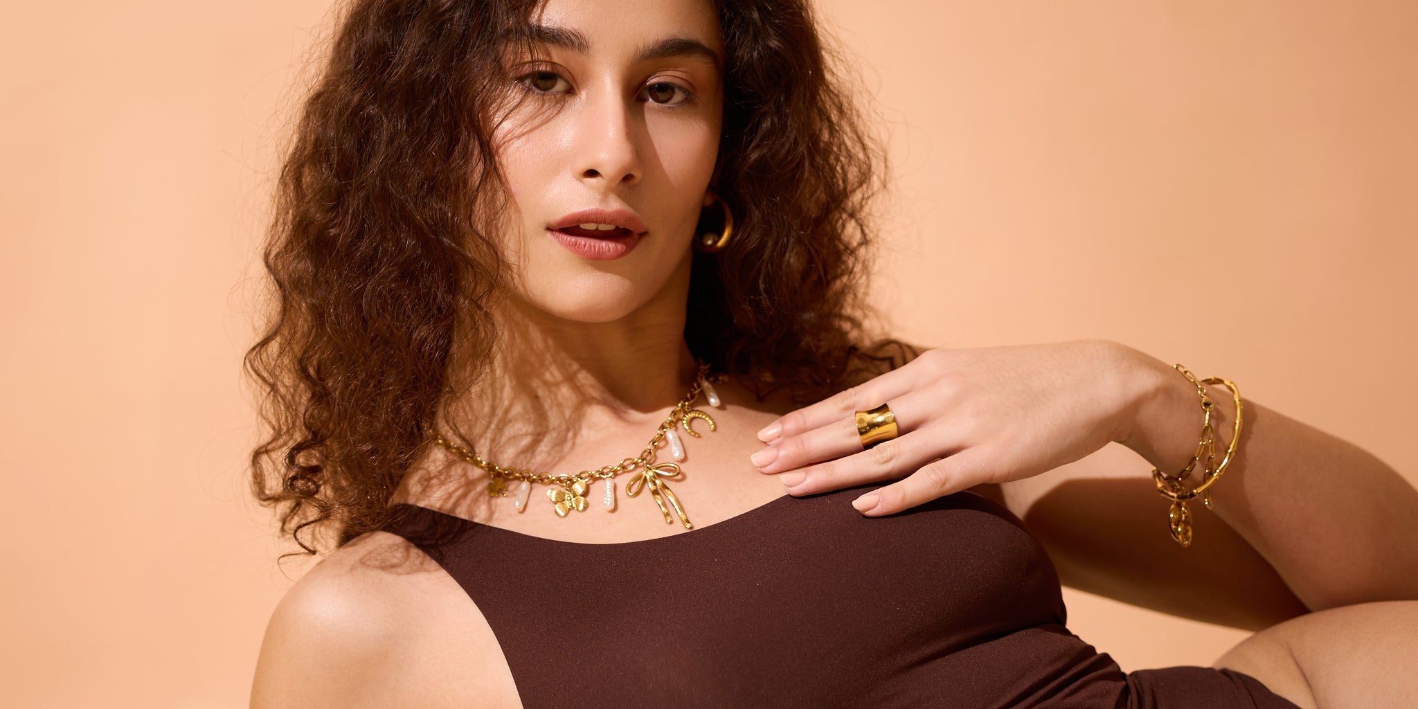 Woman wearing a layered 18K gold-plated charm necklace with pearls, bold gold ring, hoop earrings, and matching bracelets, styled with a brown sleeveless top against a warm beige background.
