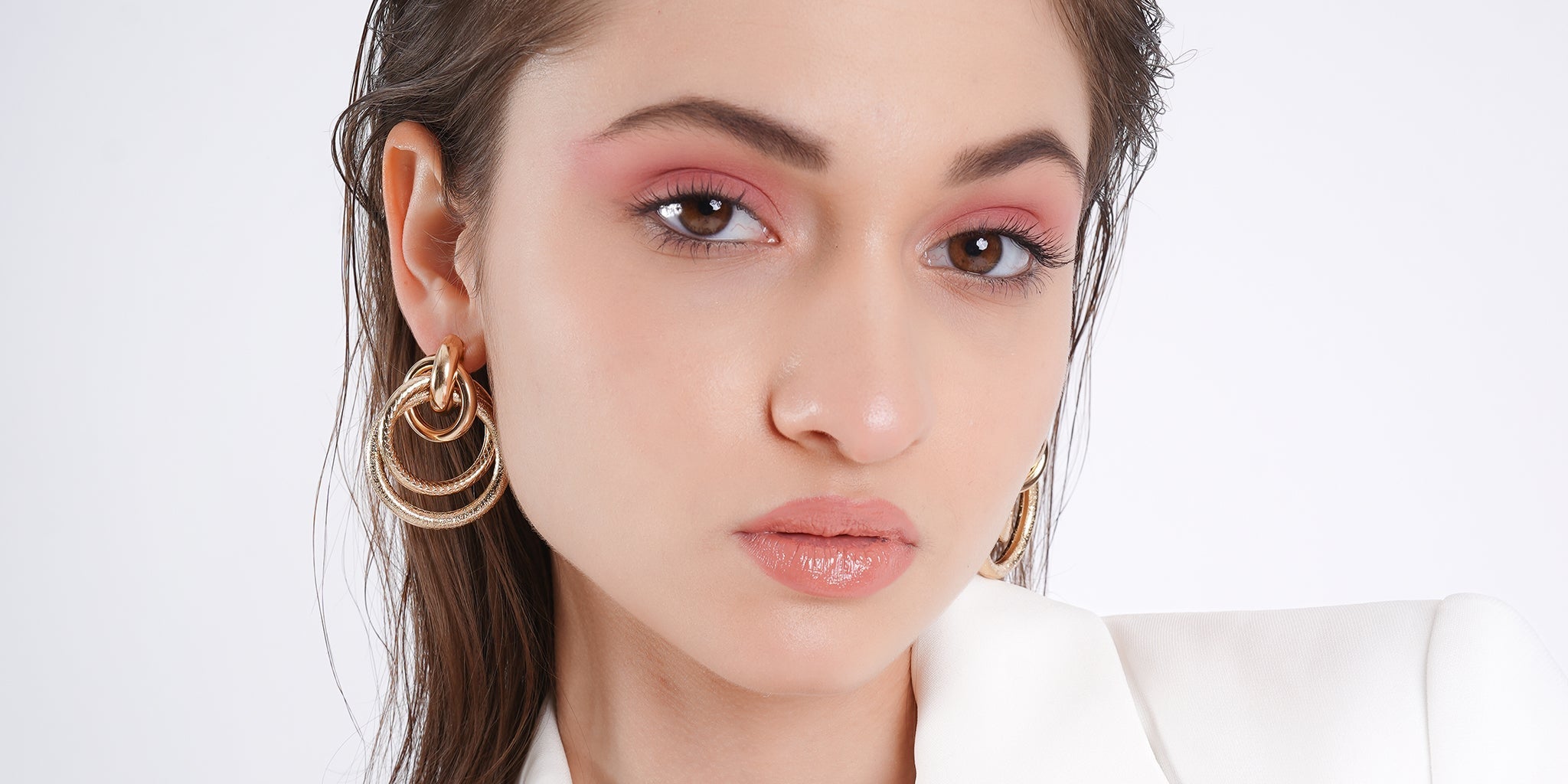 lose-up portrait of a woman wearing layered gold hoop earrings and a white outfit, styled with soft glam makeup and a confident expression.