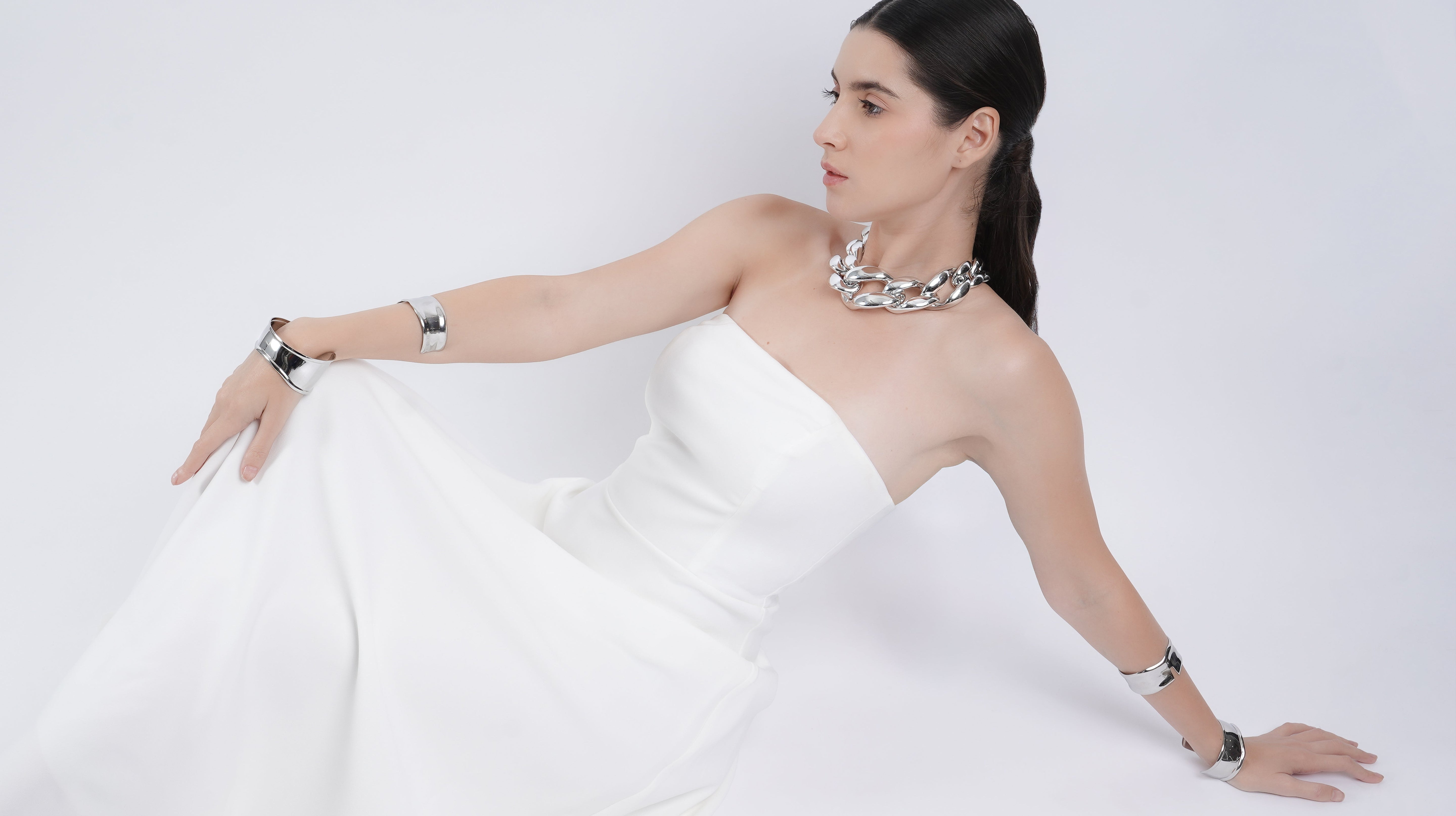 Woman in an elegant white strapless gown styled with bold 18K silver-tone statement jewelry set by Embrell, featuring a chunky necklace, bracelets, and rings; showcasing modern luxury with minimalist sophistication.