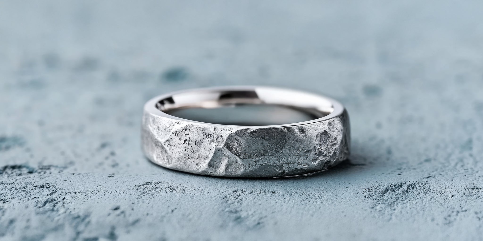 A oxidized silver ring with a hammered, textured surface is placed on a light blue, textured background.