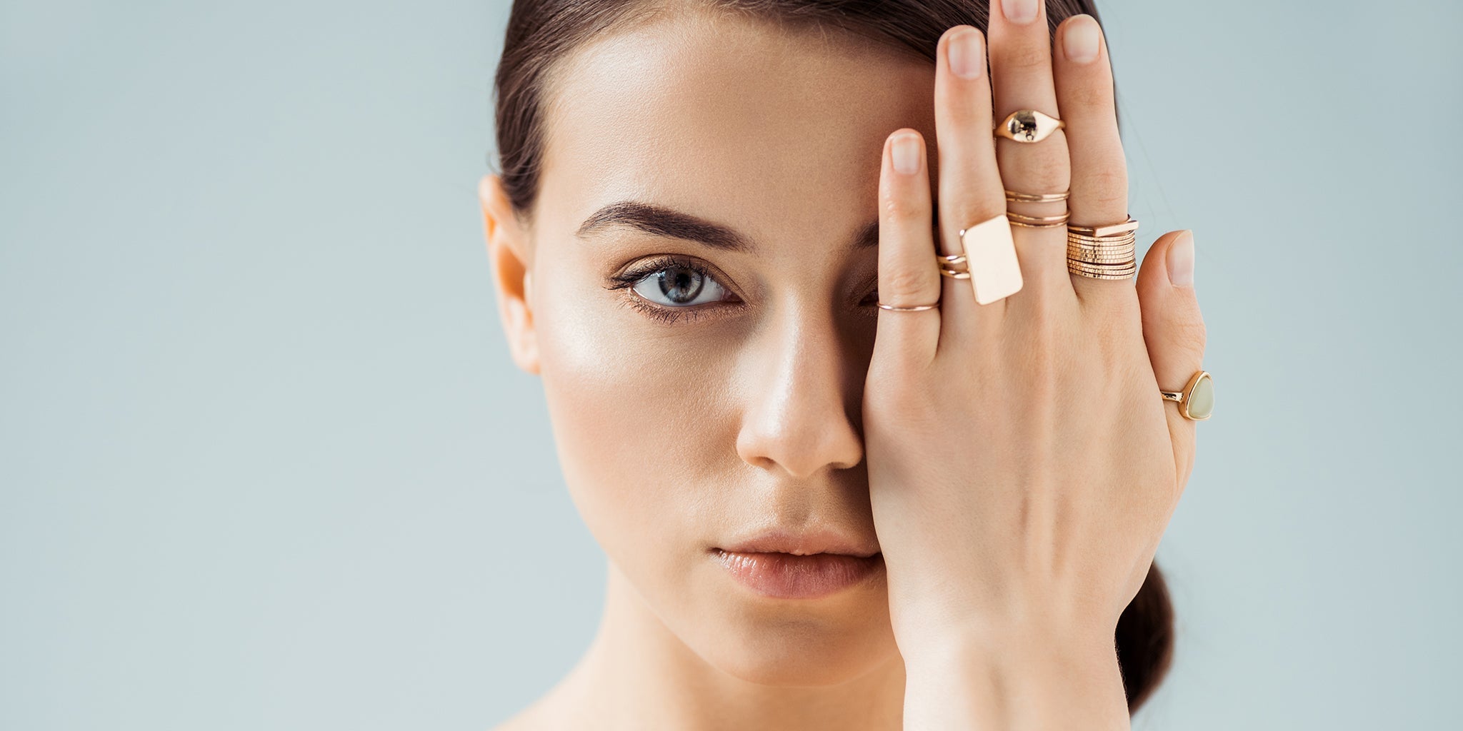 Woman wearing multiple gold rings on her fingers, covering one eye with her hand, showcasing modern and minimalist jewelry.