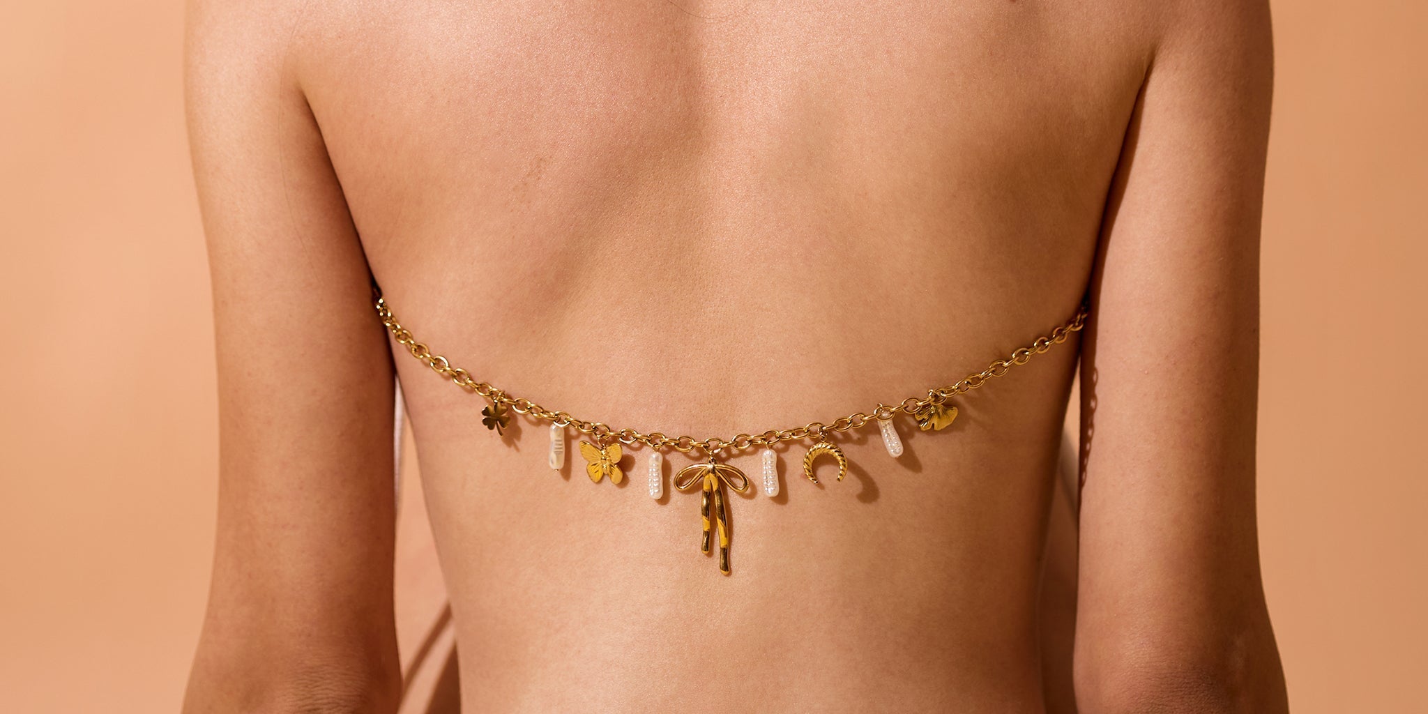 Back view of a woman wearing a delicate 18K gold-plated charm chain with mixed pendants and pearls styled across her back, set against a warm beige background.