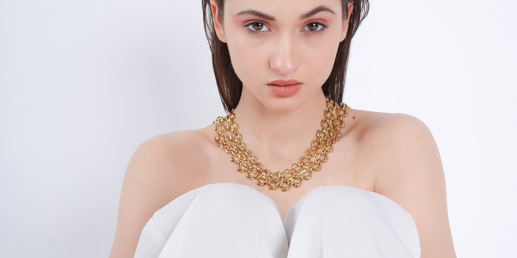Model wearing a bold 18K gold-plated statement chain necklace with interlinked oval links, styled against a white strapless outfit on a minimal background.