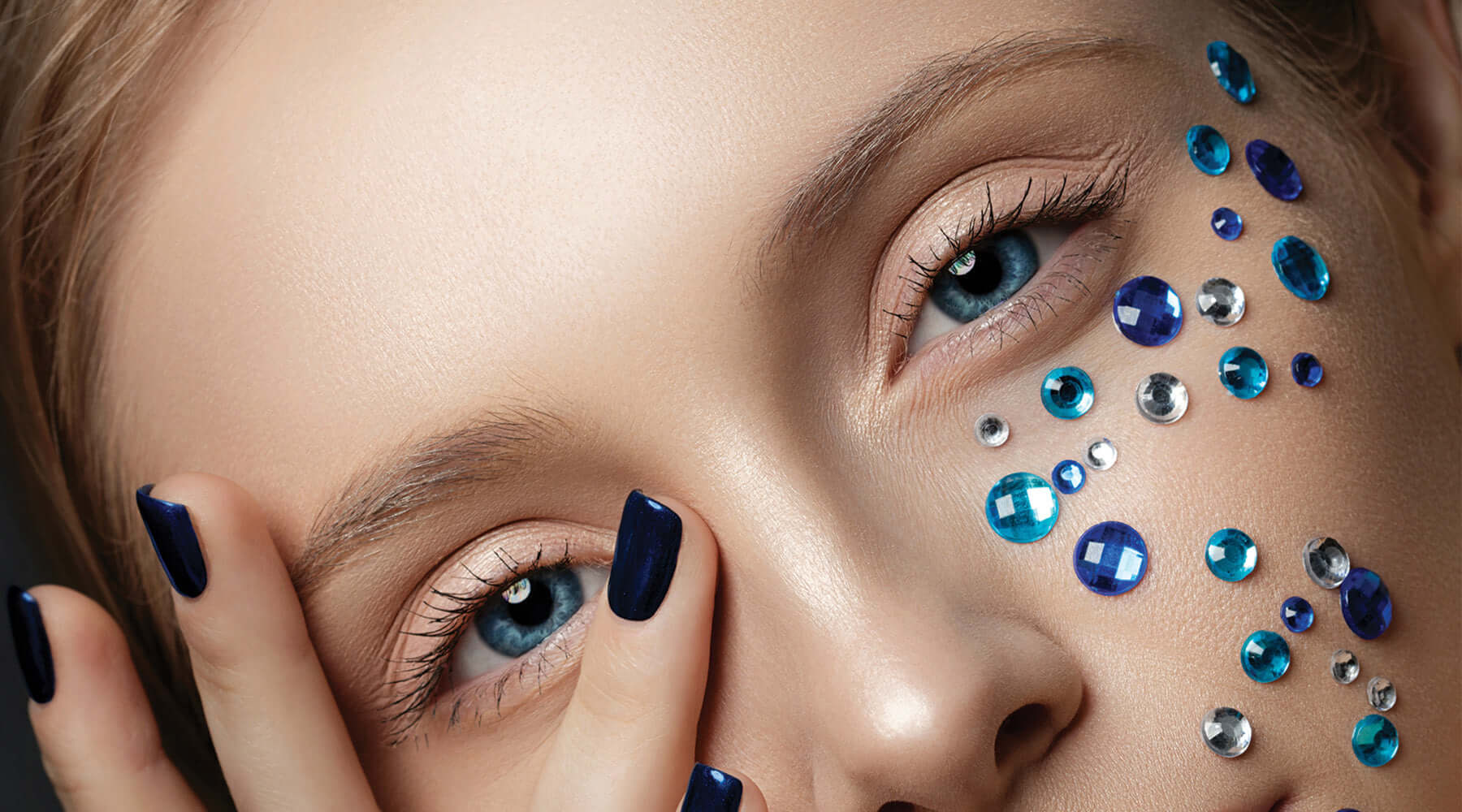 A mesmerizing close-up of bold creativity, where sparkling blue and silver gems cascade across luminous skin. Paired with deep navy nails and captivating blue eyes, this look radiates confidence and artistic elegance.