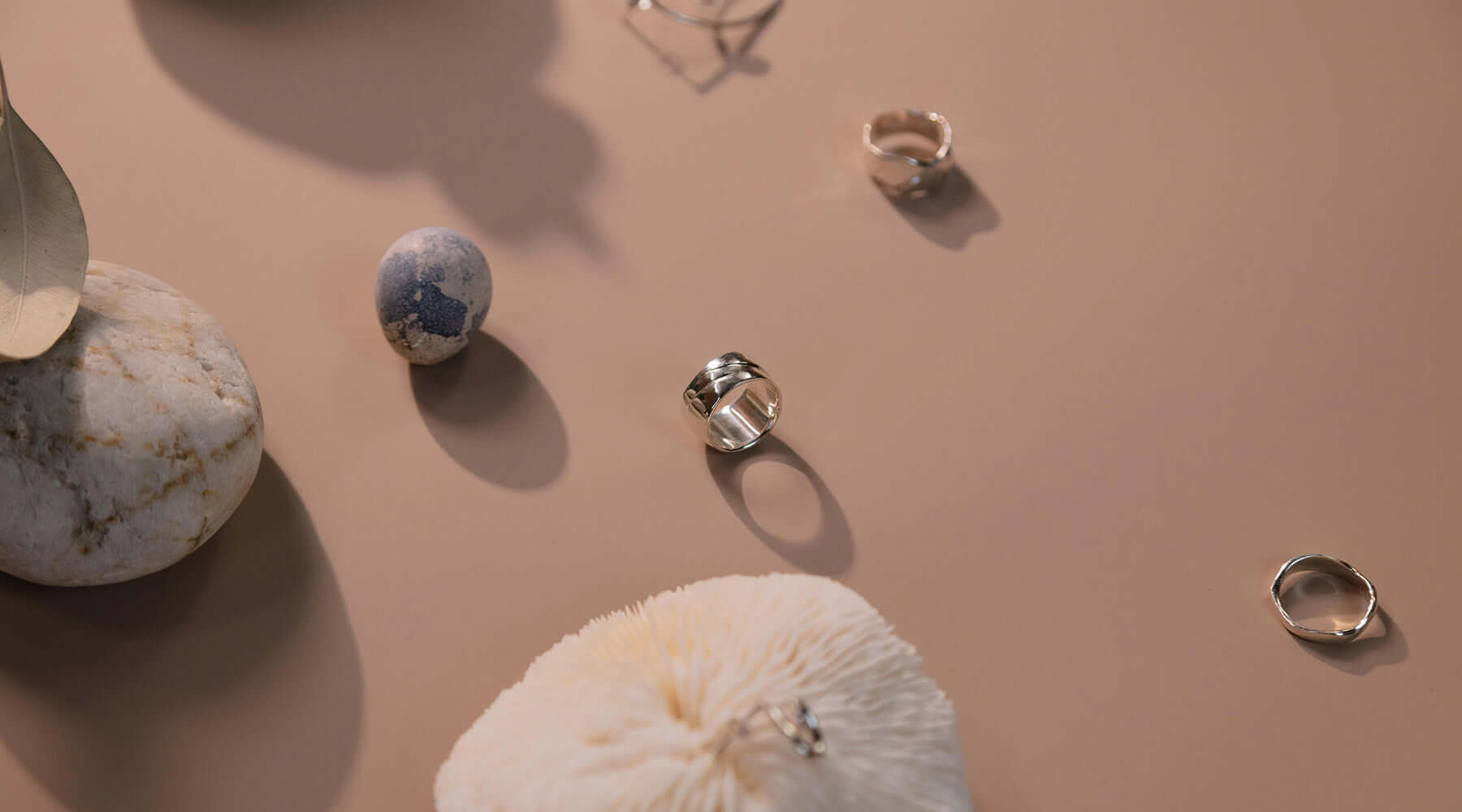 A collection of silver rings displayed on a neutral beige surface, accompanied by textured stones and decorative elements. Soft lighting and shadows enhance the elegant and minimalist aesthetic.