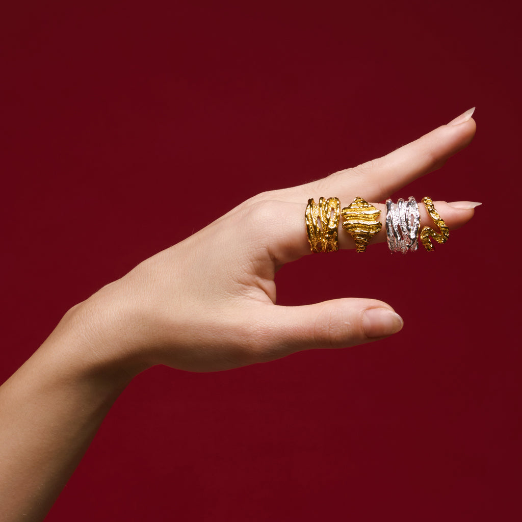 A woman in a white tank top, adorned with multiple 18K gold plated rings on her fingers, smiling softly at the camera.