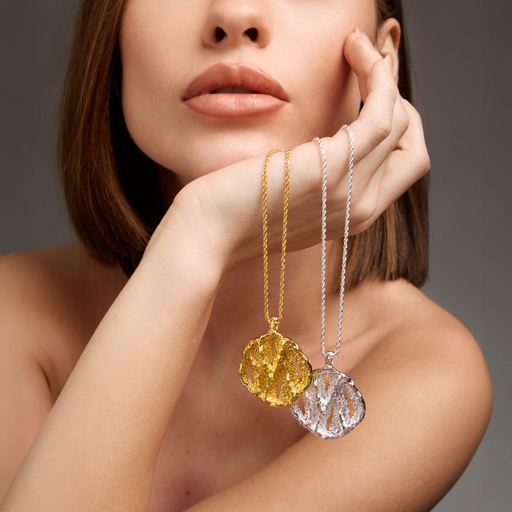 Woman holding gold and silver necklaces with circular pendants against a neutral background