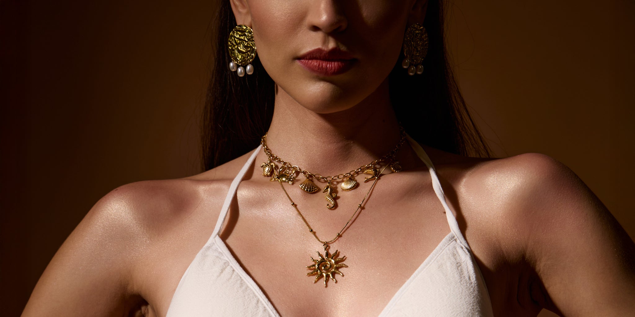 Woman wearing 18K gold plated jewellery including earrings and necklaces against a brown background