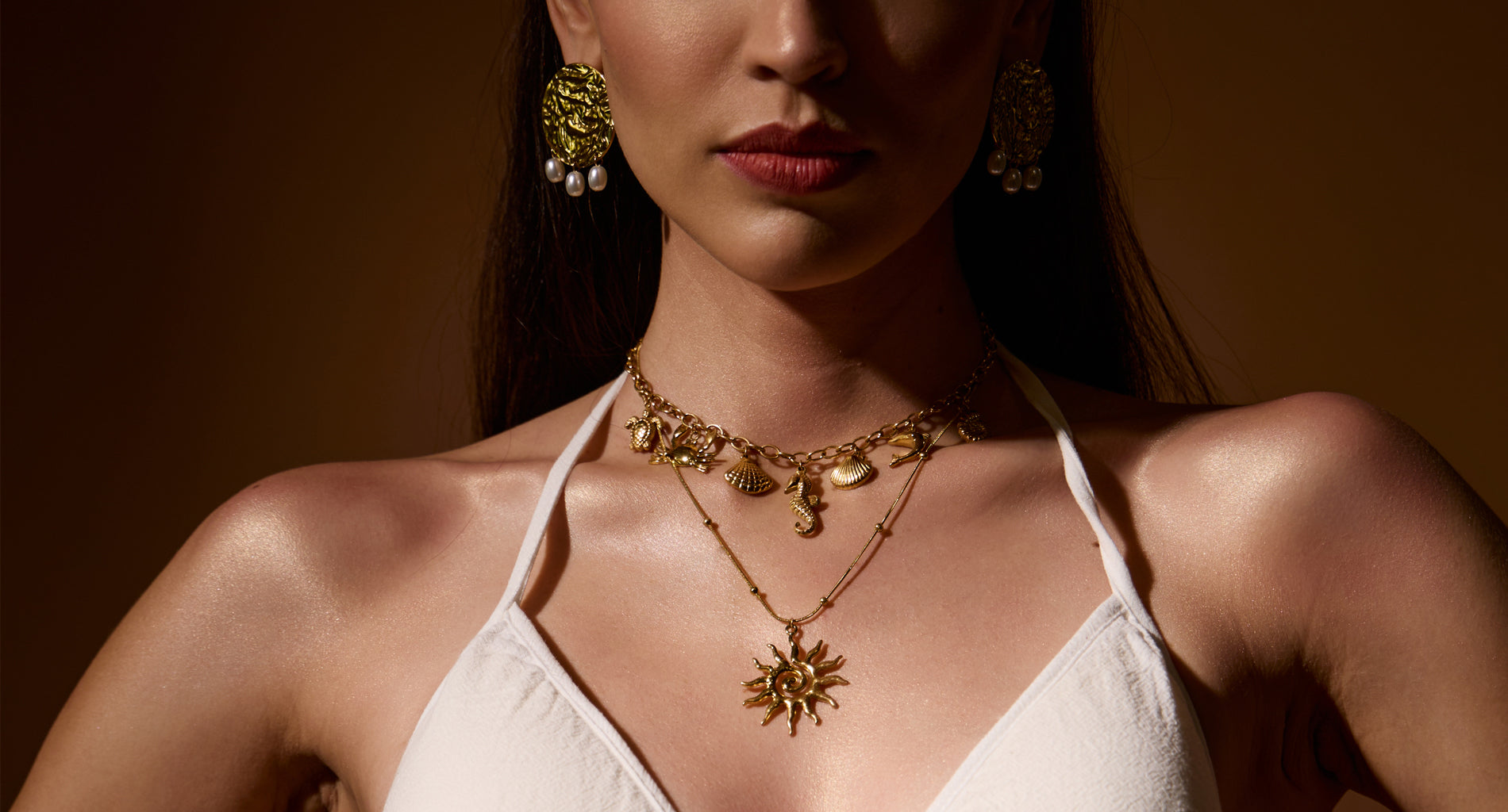 Woman wearing 18K gold plated jewellery including earrings and necklaces against a brown background