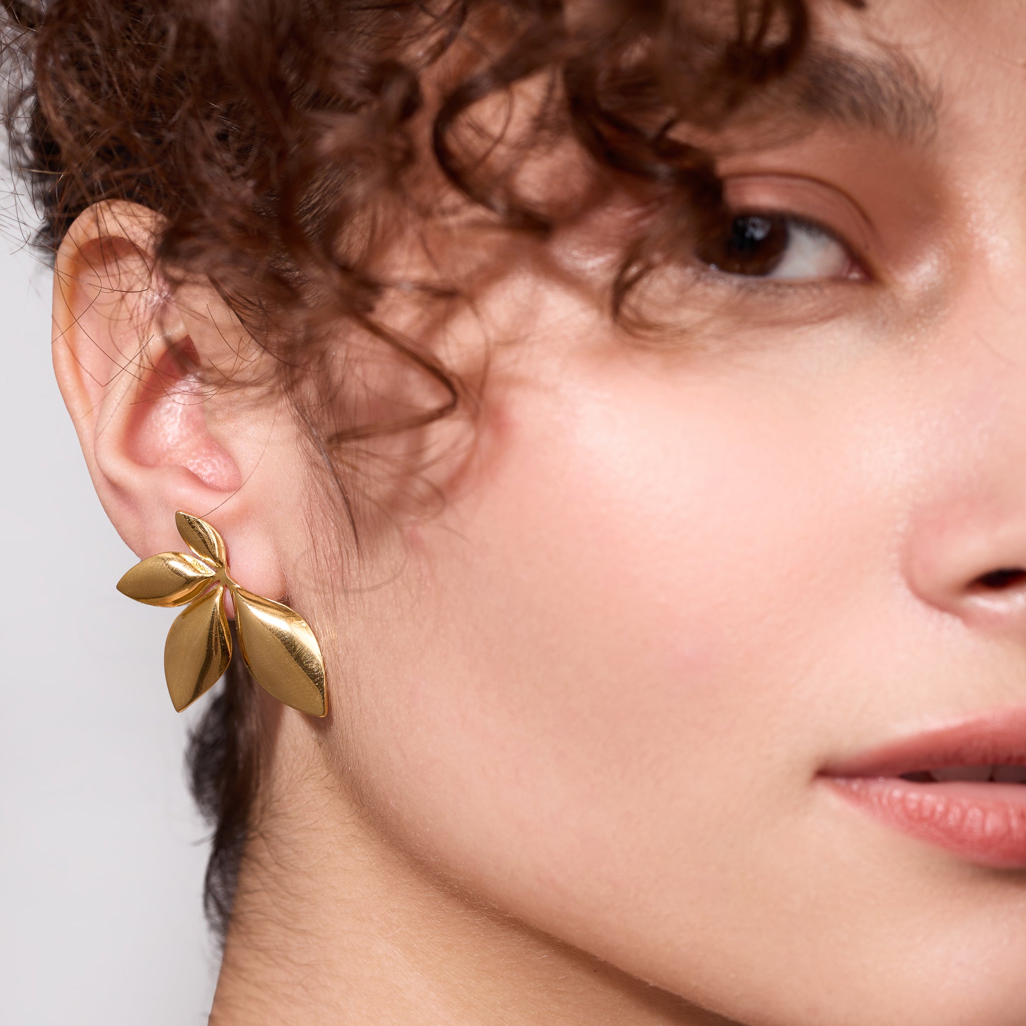 Close-up of a person wearing a 18K gold plated leaf-shaped earring against a neutral background