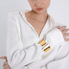 A woman in a crisp white button-down shirt showcasing a bold, polished gold cuff bracelet on her wrist, reflecting light and emphasizing its smooth, structured design.