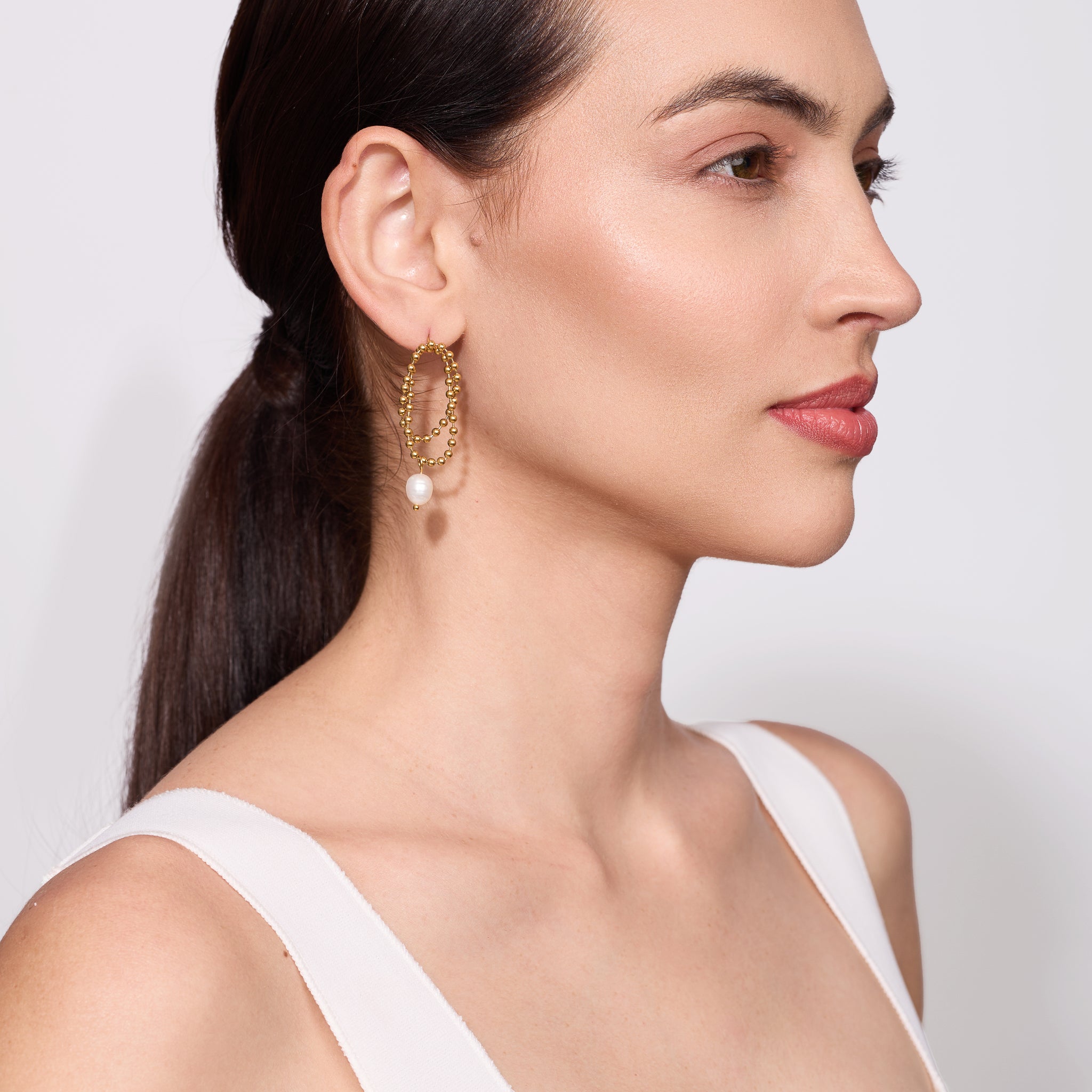 Woman wearing gold earrings with a pearl, against a plain background